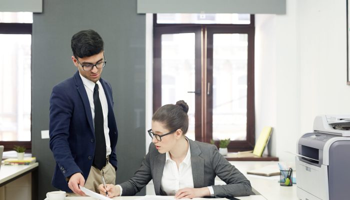 Pretty young manager making corrections in document while having informal working meeting with her handsome colleague, interior of modern open plan office on background